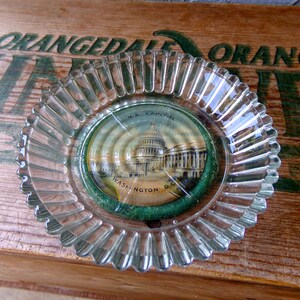 May include: A clear glass ashtray with a fluted edge. The ashtray has a round, paper sticker in the center with a picture of the US Capitol building and the text "U.S. Capitol Washington D.C."