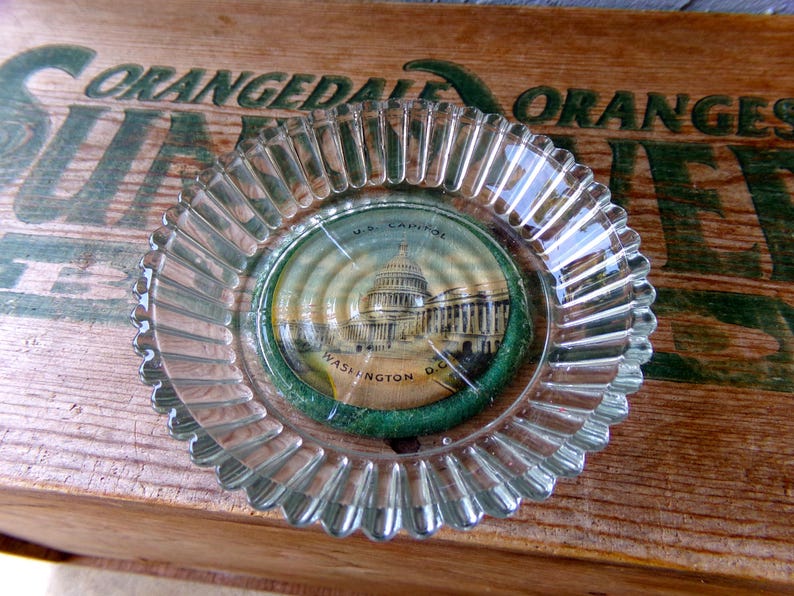 May include: A clear glass ashtray with a fluted edge. The ashtray has a round, paper sticker in the center with a picture of the US Capitol building and the text "U.S. Capitol Washington D.C."