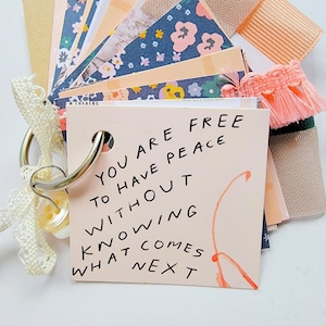 May include: A pink card with the text "You are free to have peace without knowing what comes next" written in black marker. The card is attached to a silver key ring with a white lace ribbon and a small, clear button.