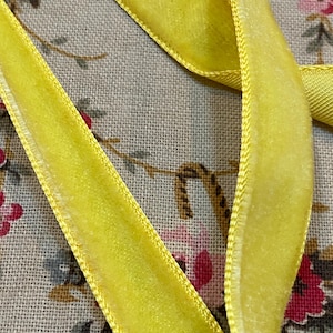 May include: Close-up of bright yellow velvet ribbon with a stitched edge. The ribbon is arranged on a floral patterned fabric background. The ribbon's texture is soft and plush, with a slight sheen.