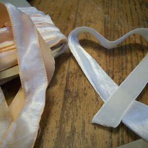 May include: Two satin ribbons, one peach and one white, are arranged on a wooden surface. The white ribbon is shaped into a heart.