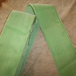May include: A green velvet ribbon with a satin edge. The ribbon is folded in half and lying on a beige mesh fabric.