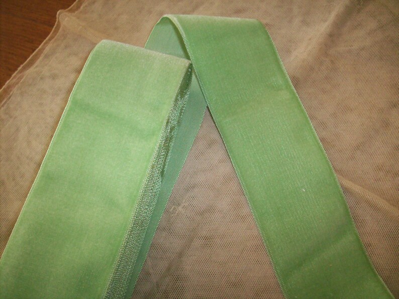 May include: A green velvet ribbon with a satin edge. The ribbon is folded in half and lying on a beige mesh fabric.