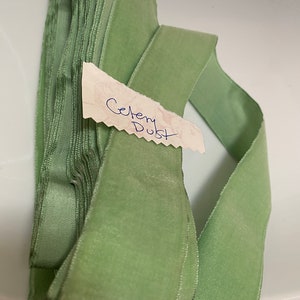May include: Celery Dust velvet ribbon, a soft, pale green hue, is neatly stacked and partially unrolled. A small tag with the handwritten words "Celery Dust" is attached. The ribbon is likely used for crafts, sewing, or gift wrapping.