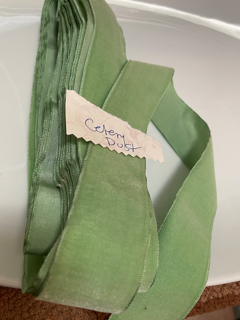 May include: Celery Dust velvet ribbon, a soft, pale green hue, is neatly stacked and partially unrolled. A small tag with the handwritten words "Celery Dust" is attached. The ribbon is likely used for crafts, sewing, or gift wrapping.