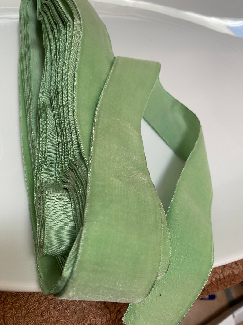 May include: A roll of soft, sage green velvet ribbon. The ribbon is folded and stacked, with a length of ribbon unrolled and visible. The ribbon has a smooth, luxurious texture, perfect for crafts and embellishments.