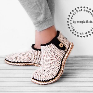 Puede incluir: Un par de zapatillas de crochet blancas y marrones con ribete negro y un botón marrón. Las zapatillas están siendo usadas por una persona de pie sobre un suelo de madera.