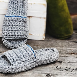 Op de afbeelding: Een paar grijze gehaakte slippers met blauwe bies. De slippers liggen op een houten oppervlak. De tekst "Crochet pattern by magic4kids" is zichtbaar in de rechterbenedenhoek van de afbeelding.