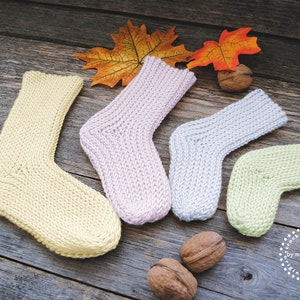 May include: Four pairs of knitted socks in different colors: yellow, light purple, light blue, and light green. The socks are arranged on a wooden surface with fall leaves and walnuts.