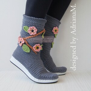 May include: Gray crochet boots with a white sole and floral embellishments. The boots are decorated with pink and green crocheted flowers and brown stems. The text "crochet boots pattern magic4kids" is visible on the boots.