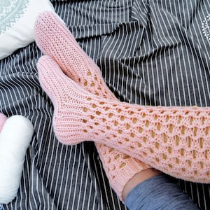 May include: A pair of pink crocheted leg warmers with a lacy pattern. The leg warmers are on a striped background.