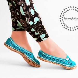 Puede incluir: Un par de zapatillas de crochet azul turquesa con ribete blanco y suela de yute marrón. Las zapatillas tienen una pequeña flor como adorno en la punta.
