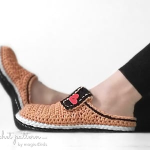 Könnte beinhalten: Gecrochete Hausschuhe in hellbrauner Farbe mit schwarzem und weißem Besatz. Die Hausschuhe haben einen herzförmigen Knopf am Riemen. Der Text "_crochet pattern_ by magic4kids" befindet sich am unteren Rand des Bildes.