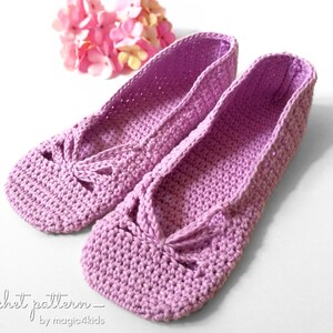 Puede incluir: Un par de zapatillas de crochet de color lavanda con un detalle de lazo. Las zapatillas están sobre un fondo blanco.