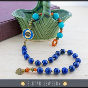 May include: A blue lapis lazuli mala prayer bead necklace with gold accents and a heart charm that says "Made with Love". The necklace is displayed on a wooden surface with a green ceramic bowl and a book.