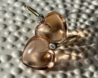 Puede incluir: Un par de pendientes en forma de corazón. Cada pendiente presenta un corazón translúcido de color melocotón con un poste dorado. Una pequeña piedra redonda y transparente está engastada en la parte superior de cada corazón. Los pendientes se muestran sobre una superficie blanca texturizada.