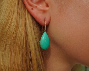 May include: A drop earring featuring a smooth, teardrop-shaped turquoise stone with subtle brown matrix inclusions. The stone hangs from a thin, gold-toned metal hook that is encrusted with a row of small, clear, sparkling pavé-set stones.