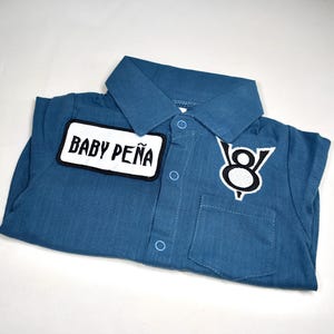 May include: A blue collared shirt with a button-down front, featuring a white rectangular patch with the black text BABY PE&Ntilde;A on the right side and a black and white V8 engine logo patch on the left side above a small chest pocket.