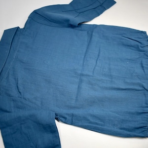 May include: A solid blue long-sleeved shirt featuring a classic collar, a button-down front placket, and a rounded hemline. The garment is crafted from a textured fabric and displays a simple, functional design with cuffed sleeves and visible stitching.