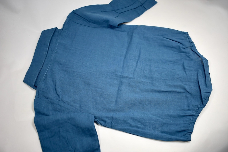 May include: A solid blue long-sleeved shirt featuring a classic collar, a button-down front placket, and a rounded hemline. The garment is crafted from a textured fabric and displays a simple, functional design with cuffed sleeves and visible stitching.