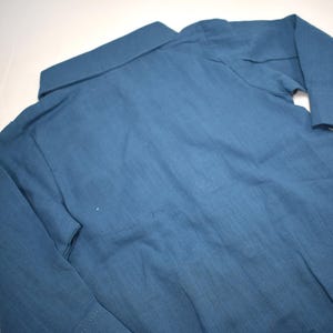 May include: A long-sleeved button-down shirt in a solid muted blue color. The garment features a standard collar, a front button placket, and cuffs on the sleeves. The fabric has a visible woven texture, and the shirt is laid flat for display.