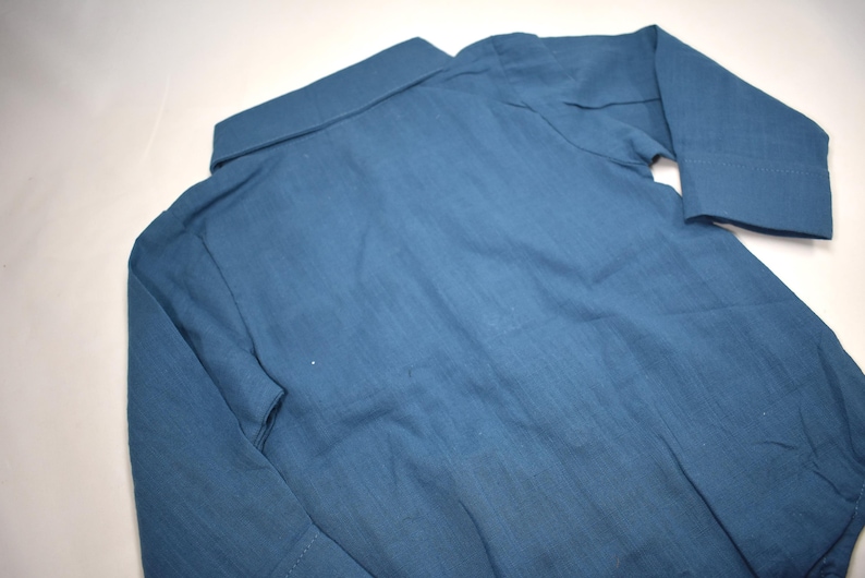 May include: A long-sleeved button-down shirt in a solid muted blue color. The garment features a standard collar, a front button placket, and cuffs on the sleeves. The fabric has a visible woven texture, and the shirt is laid flat for display.