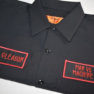 Peut inclure: Chemise de travail noire avec deux patchs brodés rouges. Le patch de gauche dit "T. Gleason" et le patch de droite dit "Man vs Machine".