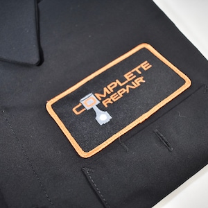 Puede incluir: Una camisa negra de botones con un parche rectangular en el pecho. El parche tiene un borde naranja y muestra el texto COMPLETE REPAIR en naranja, con un gr&aacute;fico de un pist&oacute;n de motor gris integrado en la letra O.