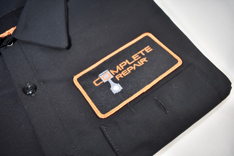 Puede incluir: Una camisa negra de botones con un parche rectangular en el pecho. El parche tiene un borde naranja y muestra el texto COMPLETE REPAIR en naranja, con un gr&aacute;fico de un pist&oacute;n de motor gris integrado en la letra O.