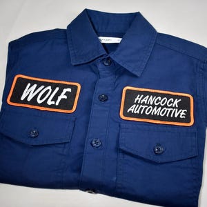 May include: A dark blue button-down shirt featuring two chest pockets and two rectangular patches with orange borders. The left patch displays the text WOLF in white, and the right patch displays the text HANCOCK AUTOMOTIVE in white lettering.