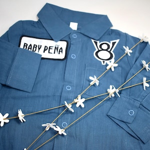 May include: A blue long-sleeved button-down shirt featuring a white rectangular patch with the black text BABY PE&Ntilde;A and a white and black V8 engine emblem patch on the chest. The shirt includes a collar, a front pocket, and buttoned cuffs.
