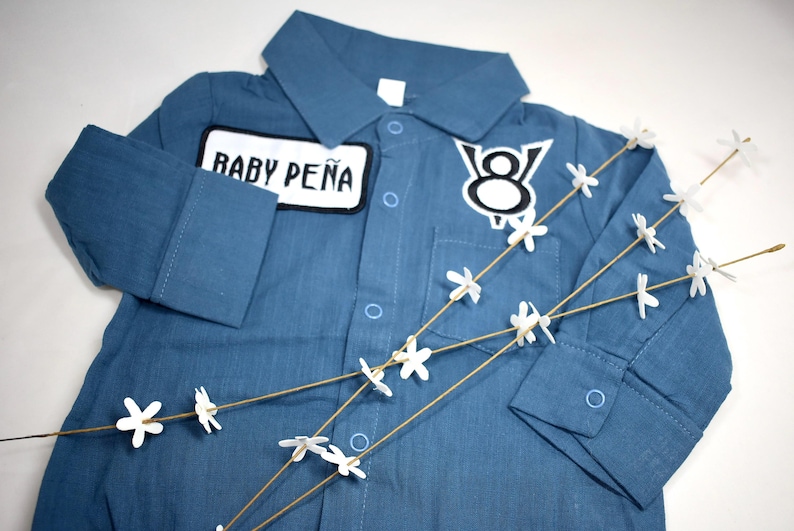 May include: A blue long-sleeved button-down shirt featuring a white rectangular patch with the black text BABY PE&Ntilde;A and a white and black V8 engine emblem patch on the chest. The shirt includes a collar, a front pocket, and buttoned cuffs.