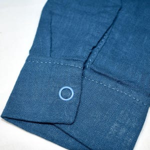 May include: A long-sleeved shirt cuff in a deep blue fabric with a visible woven texture. The cuff features a single light blue circular button centered on the flap, with matching blue stitching along the edges of the cuff and the sleeve placket.