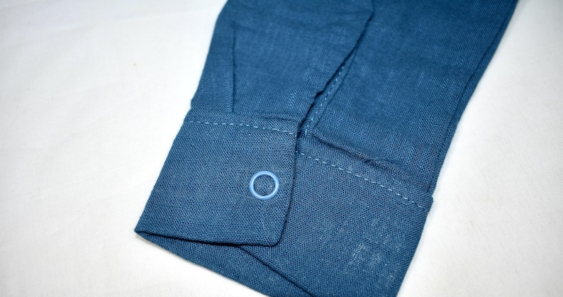 May include: A long-sleeved shirt cuff in a deep blue fabric with a visible woven texture. The cuff features a single light blue circular button centered on the flap, with matching blue stitching along the edges of the cuff and the sleeve placket.