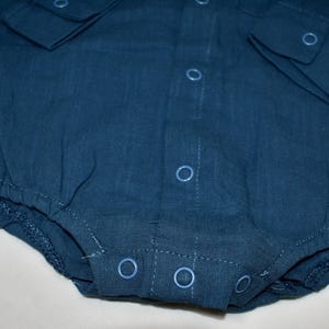 May include: A dark blue long-sleeved bodysuit made of a textured fabric. The garment features a front button placket with matching blue circular snap closures, a collar, and elasticized leg openings for a secure fit around the lower torso.
