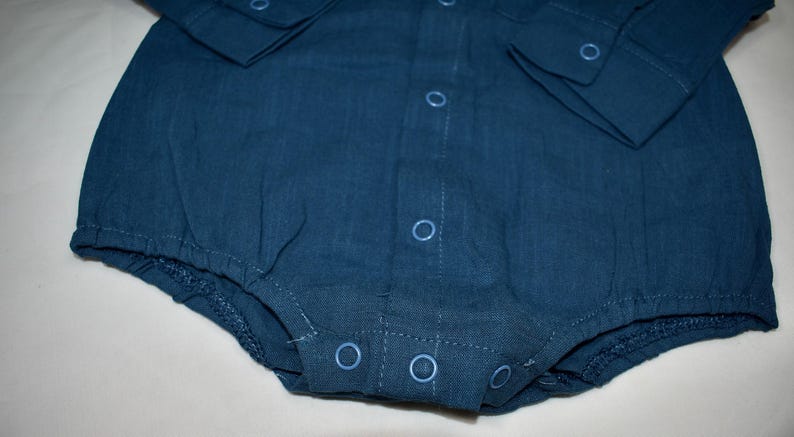 May include: A dark blue long-sleeved bodysuit made of a textured fabric. The garment features a front button placket with matching blue circular snap closures, a collar, and elasticized leg openings for a secure fit around the lower torso.