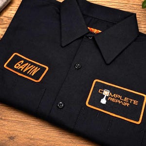May include: A black button-down work shirt featuring two embroidered patches with orange borders. The left chest patch displays the name GAVIN in orange text, while the right chest patch features the text COMPLETE REPAIR with a piston icon.