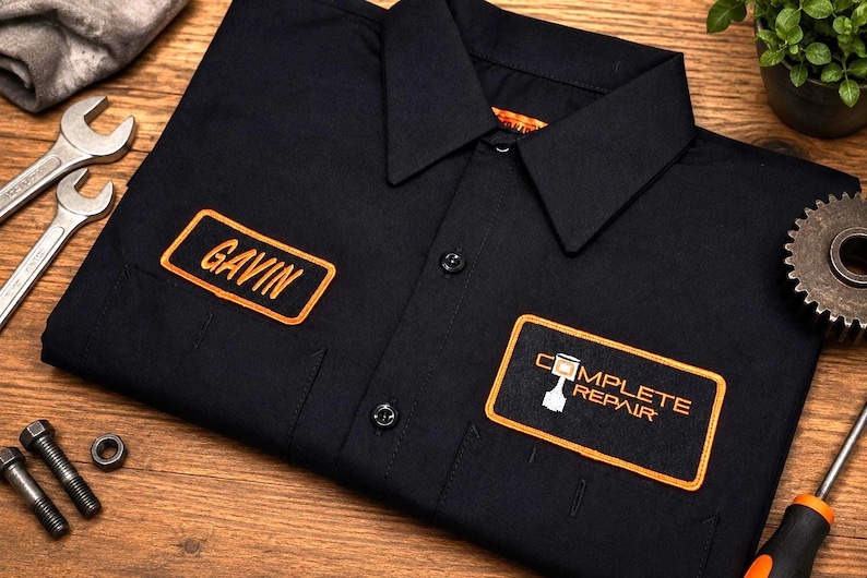 Puede incluir: Una camisa de trabajo negra con botones y dos parches bordados con bordes naranjas. El parche del pecho izquierdo muestra el nombre GAVIN en letras naranjas, mientras que el derecho tiene el texto COMPLETE REPAIR con un icono de pist&oacute;n.