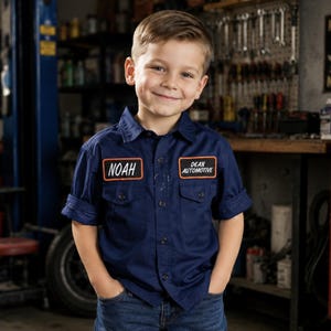 May include: A navy blue button-down work shirt with rolled sleeves, featuring two chest pockets and two rectangular patches. The left patch reads NOAH in white text, and the right patch reads DEAN AUTOMOTIVE in white text, both with orange borders.