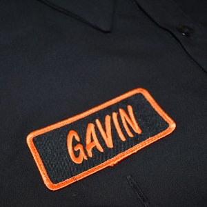 Puede incluir: Una camisa de trabajo negra con botones y un parche rectangular bordado en el pecho con el nombre GAVIN cosido en hilo naranja. El parche tiene un borde naranja grueso. Un segundo parche parcial es visible en la esquina superior derecha.