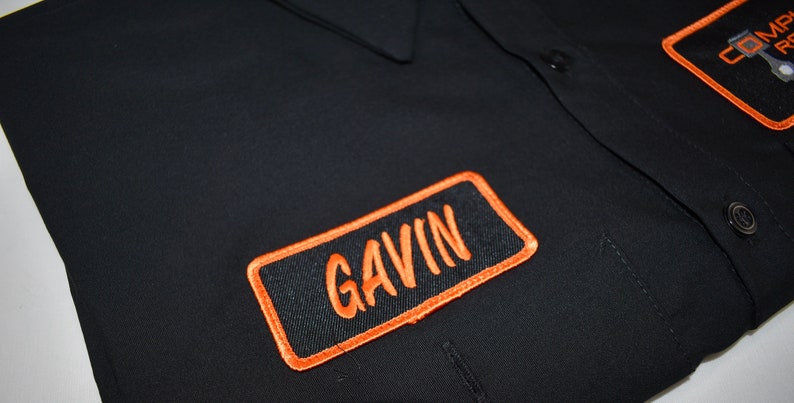 Puede incluir: Una camisa de trabajo negra con botones y un parche rectangular bordado en el pecho con el nombre GAVIN cosido en hilo naranja. El parche tiene un borde naranja grueso. Un segundo parche parcial es visible en la esquina superior derecha.