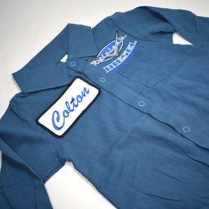 May include: A blue long-sleeve button-down shirt featuring a white rectangular patch with the name Colton embroidered in blue script. The shirt includes a chest pocket with a blue and white Diesel logo patch and a classic collar design.