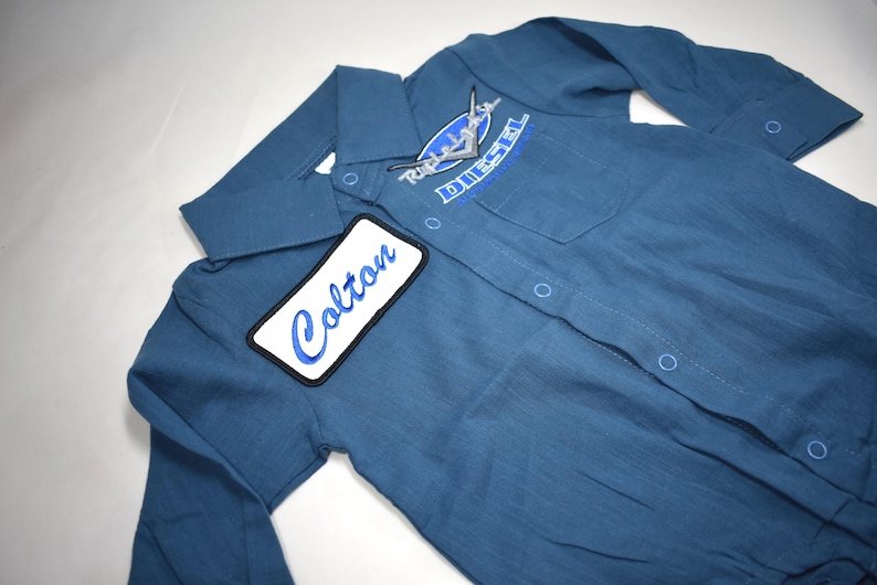 May include: A blue long-sleeve button-down shirt featuring a white rectangular patch with the name Colton embroidered in blue script. The shirt includes a chest pocket with a blue and white Diesel logo patch and a classic collar design.