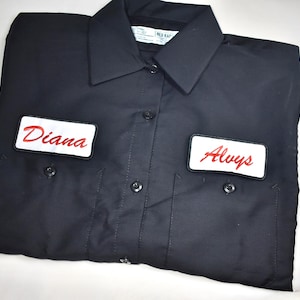 May include: Black work shirt with two chest pockets and a collar. The shirt has two patches with red embroidered text: "Diana" on the left and "Always" on the right. The shirt is folded neatly on a white surface.