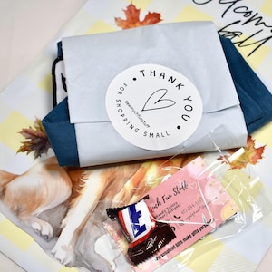 May include: A blue fabric item wrapped in white paper with a round sticker that reads Thank You For Shopping Small and a heart icon. Below is a small clear plastic bag containing a pink business card and a miniature chocolate candy bar.