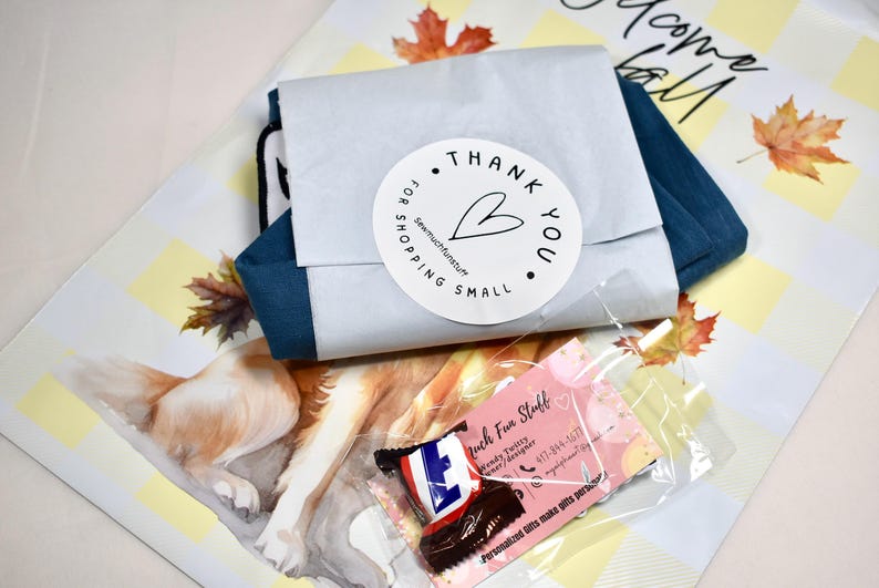 May include: A blue fabric item wrapped in white paper with a round sticker that reads Thank You For Shopping Small and a heart icon. Below is a small clear plastic bag containing a pink business card and a miniature chocolate candy bar.