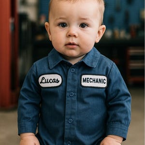 May include: A baby wearing a blue mechanic-style jumpsuit. The jumpsuit has a collared neckline, button closures, and patches that read "Lucas" and "MECHANIC". The baby is looking directly at the camera.