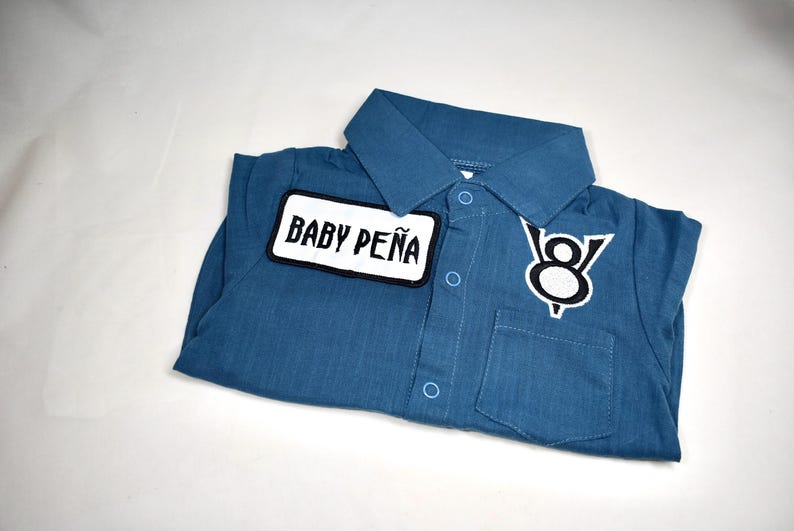 May include: A blue collared shirt with a button-down front, featuring a white rectangular patch with the black text BABY PE&Ntilde;A on the right side and a black and white V8 engine logo patch on the left side above a small chest pocket.