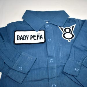 May include: A long-sleeved blue button-down shirt featuring a white rectangular patch with the black text BABY PE&Ntilde;A and a white V8 engine logo patch on the chest. The shirt includes a classic collar, a front pocket, and light blue button closures.