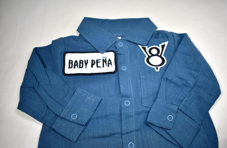 May include: A long-sleeved blue button-down shirt featuring a white rectangular patch with the black text BABY PE&Ntilde;A and a white V8 engine logo patch on the chest. The shirt includes a classic collar, a front pocket, and light blue button closures.